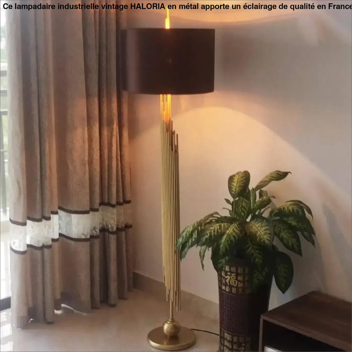 Lampadaire industrielle vintage haloria avec abat-jour cylindrique noir et base en tiges dorées émettant une lumière chaude