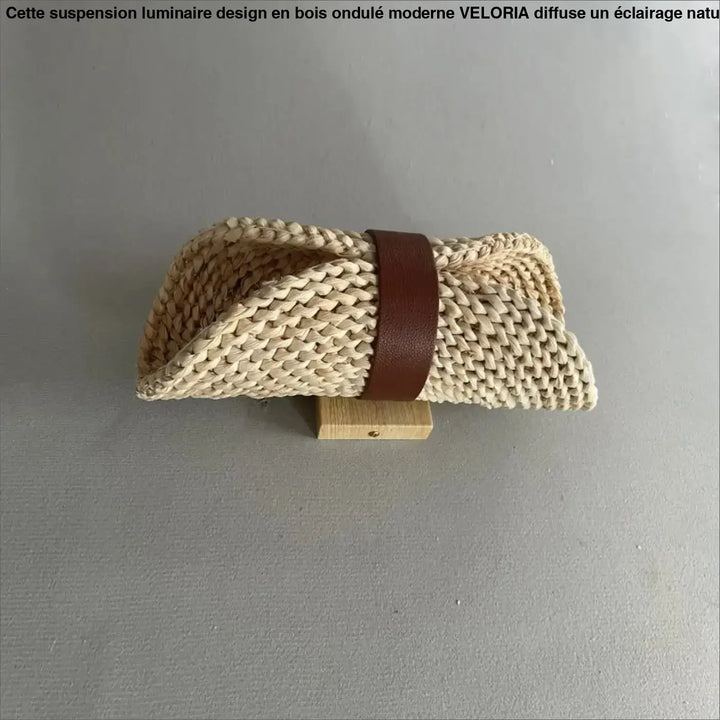 Beige woven straw clutch with brown leather strap displayed with suspension luminaire design bois ondule moderne by veloria